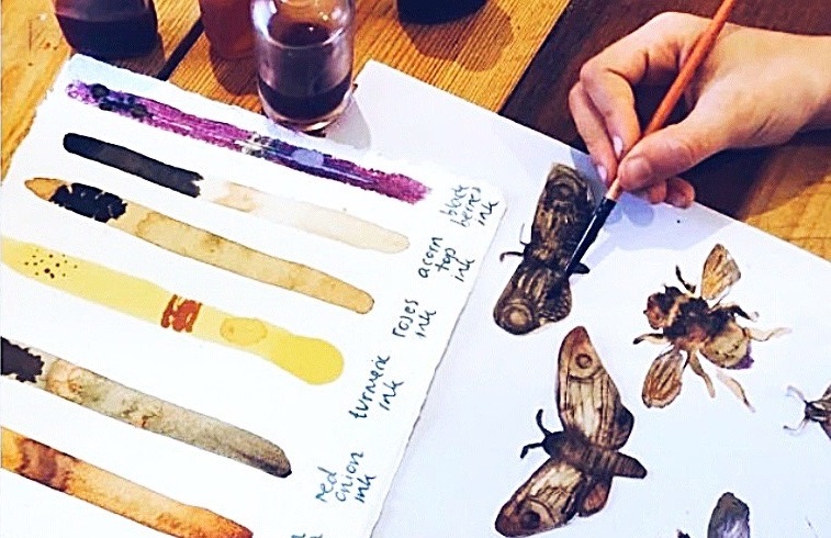 A close-up photo of an artist painting insects with watercolours. A handmade swatch of ink colours sits on the table.