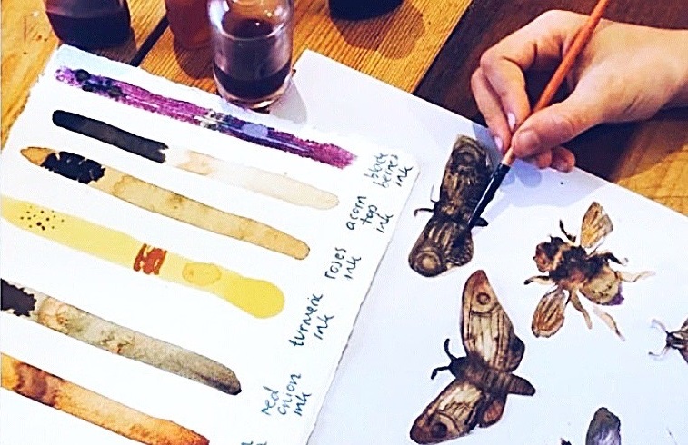 thumbnail_image001.jpg A close-up photo of an artist painting insects with watercolours. A handmade swatch of ink colours sits on the table.