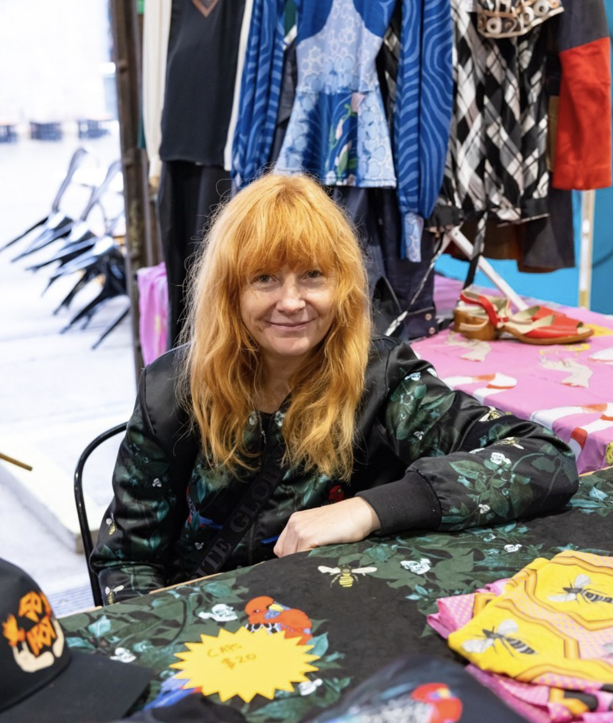 Zanny Begg sits in her studio, colourful fabric on the table in front of her. She is smiling. 